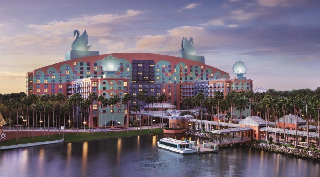 Wide shot of a large resort hotel with two huge swan sculptures on top of it next to a lake with a boardwalk and a ferry boat